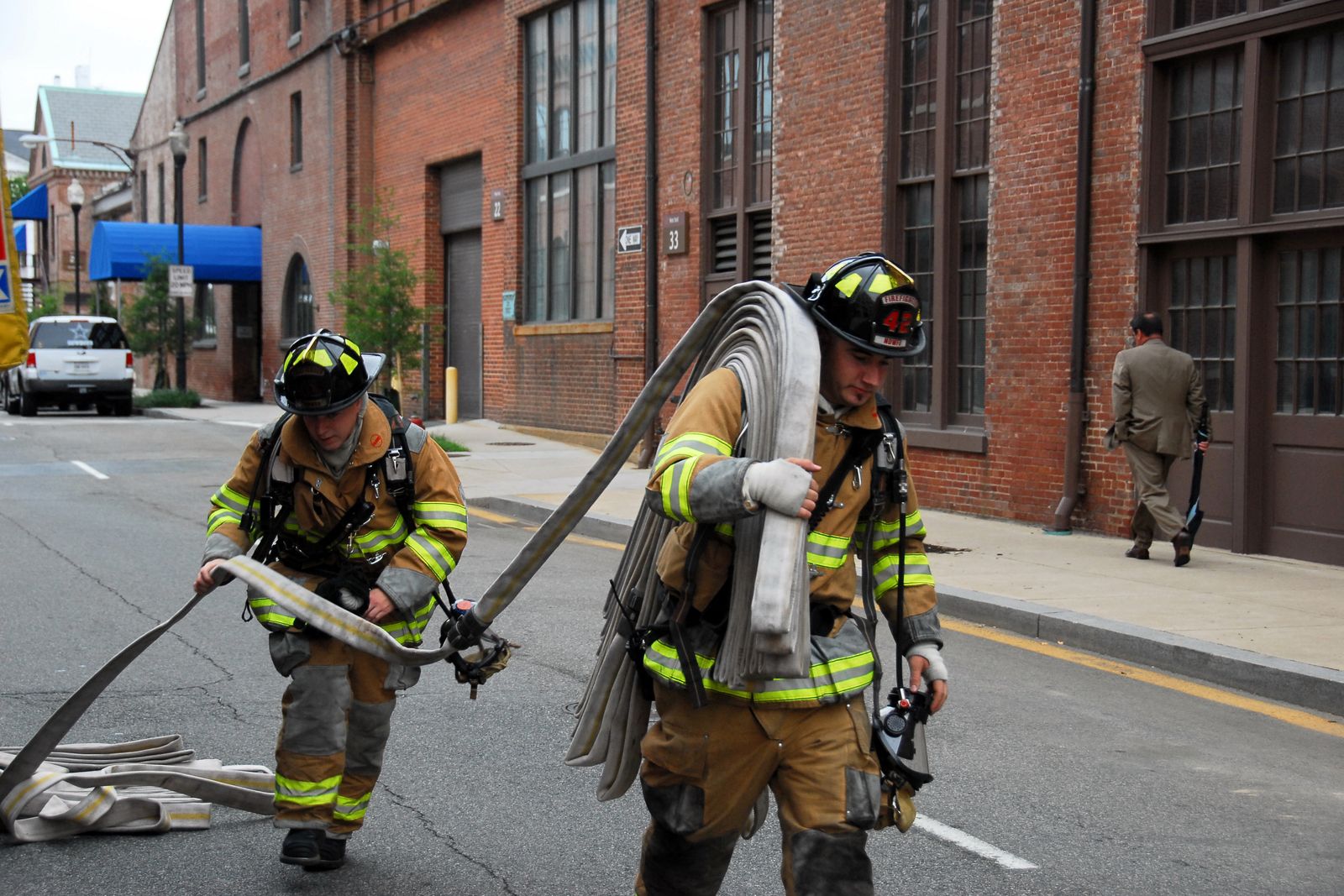 Two firefighters in turnout gear walking down a city street, one carrying a large coil of supply hose over his shoulder